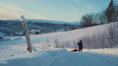 Aufnahme-Eines-Kleinen-Jungen,-Der-Den-Schneebedeckten-Hang-Entlang-Zurückgeht-Und-Tagsüber-Mit-Einem-Roten-Plastikschaufelschlitten-In-Beitostolen,-Einem-Dorf-In-Der-Grafschaft-Innlandet,-Valdres,-Beito,-Norwegen,-Schlitten-Fährt