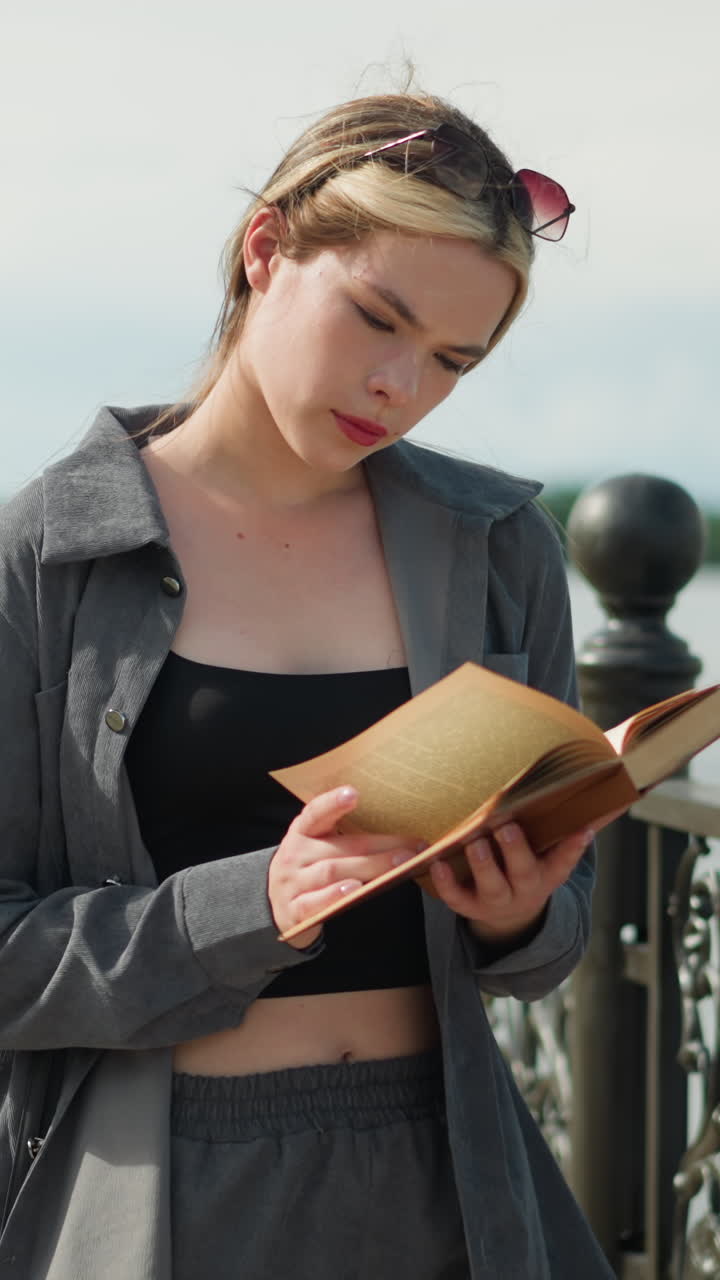 mujer con ropa gris y bolso negro se encuentra junto a una valla, volviendo a una nueva página en un libro mientras se centra, el viento sopla a través de las páginas con árboles visibles en la distancia por la orilla del río