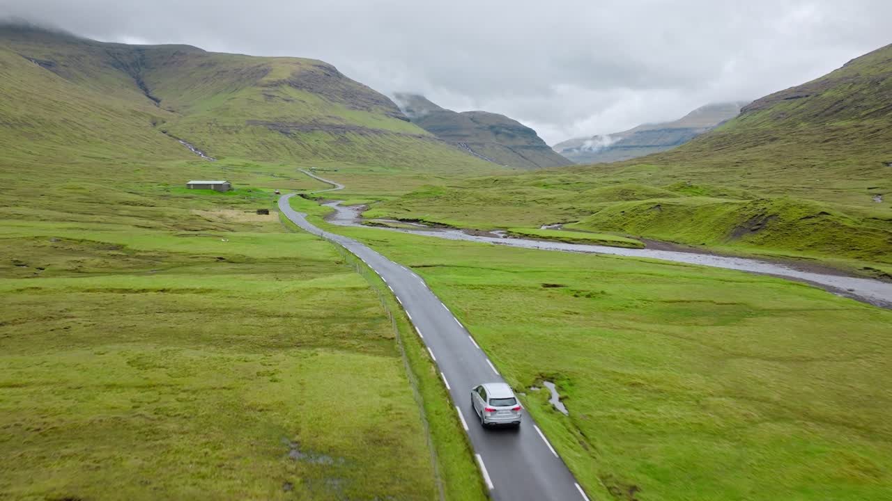 A car drives on a winding road in the Faroe Islands, surrounded by green hills