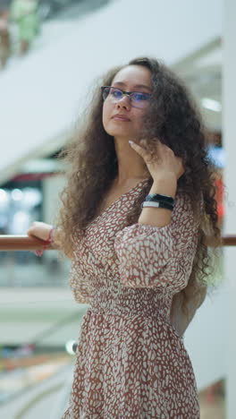 woman in vintage dress stands near iron railing, adjusting her hair thoughtfully in a bright, spacious mall, background blur shows people moving on escalator