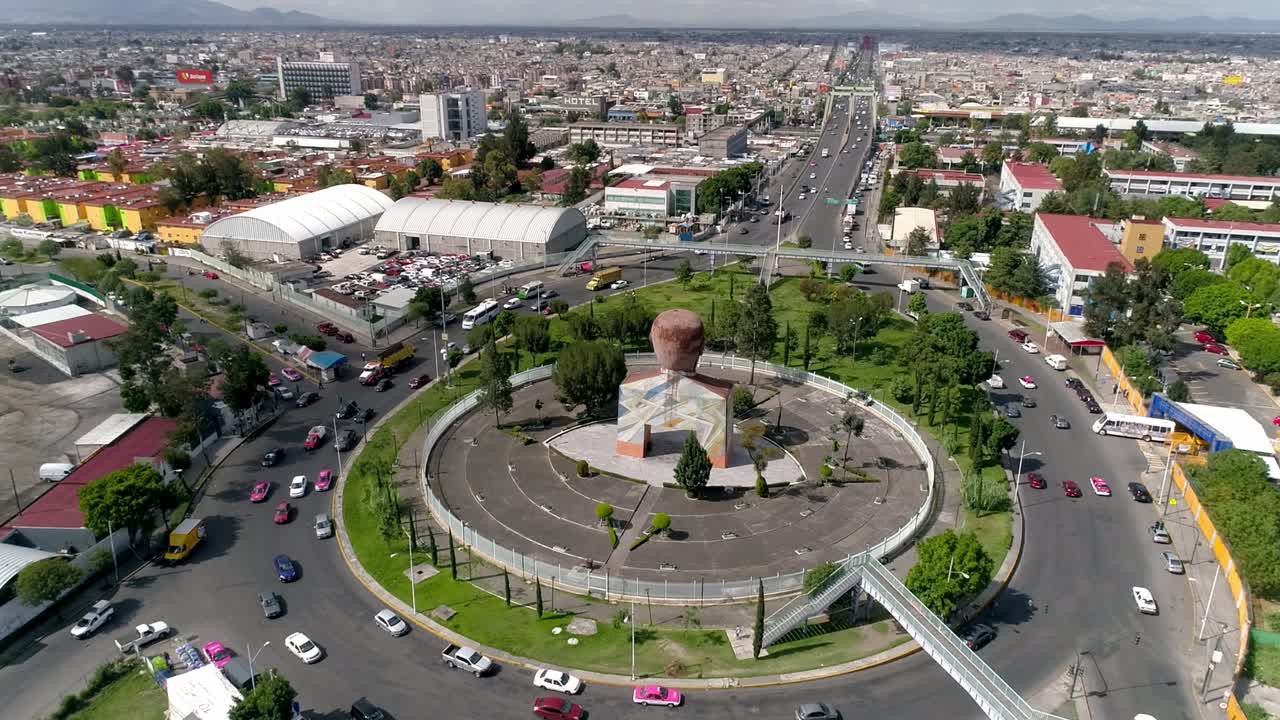 Tomada aérea de drone de uma rotatória com monumento ao centro, veículos se movendo por estradas circulares e uma rodovia se estendendo pela cidade, mostrando o fluxo de tráfego e o layout urbano.