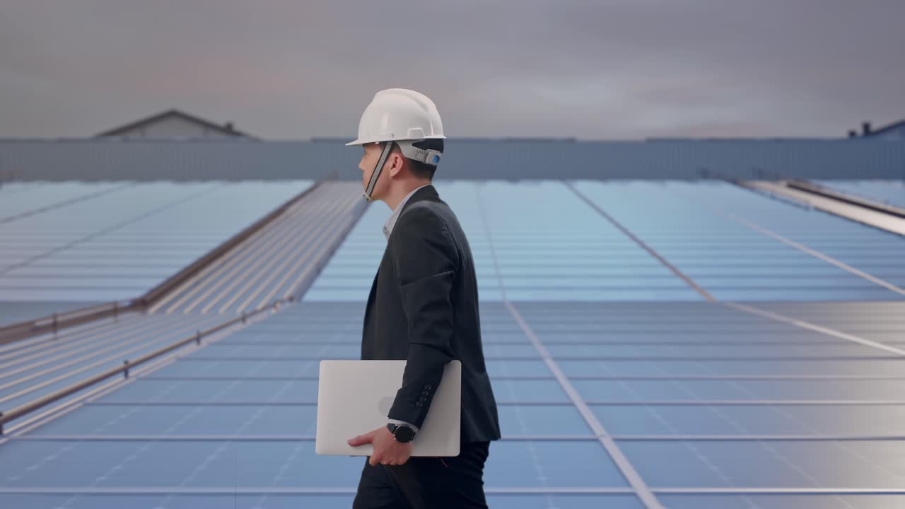 Vista laterale di uomo d'affari asiatico che tiene un computer mentre cammina su un tetto con pannelli solari industriali.