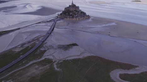 Mont-Saint-Michel-at-low-tide-with-shuttlebus-drive-on-road,-aerial-tilt-up