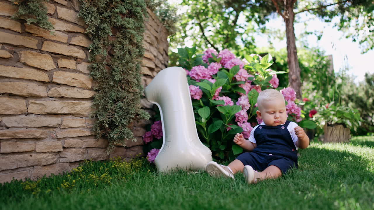 Cute baby sitting on the green grass plays with flower petals. Balloon of number one shape is beside the kid. First birthday concept.
