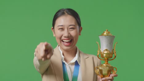 close up of asian business woman in a suit with a gold medal and trophy touching her chest then smiling and pointing to camera being proud of herself on green screen background in the studio