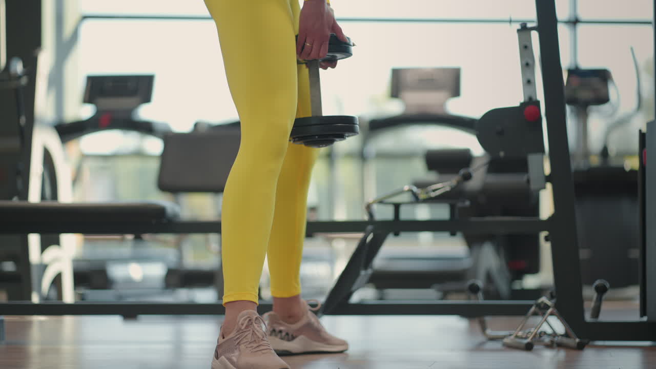 Young Hispanic brunette woman squats holding a dumbbell in the gym in a yellow tracksuit