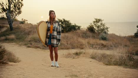 una chica rubia con una camisa a cuadros está de pie en una pendiente cerca del mar y sostiene una tabla de surf de madera en sus manos contra el amanecer de la mañana. una chica está de pé en una pista cerca del mar, inspecciona el lugar y espera a alguien para surfear en el mar