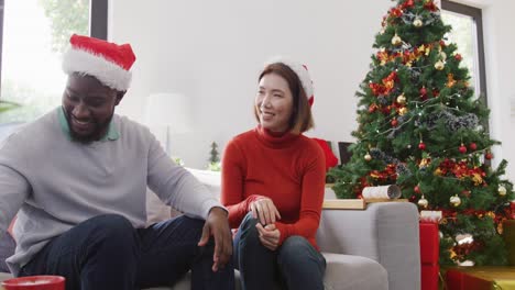 Video-of-happy-diverse-couple-in-santa-hats-exchanging-christmas-gift-and-smiling-at-home