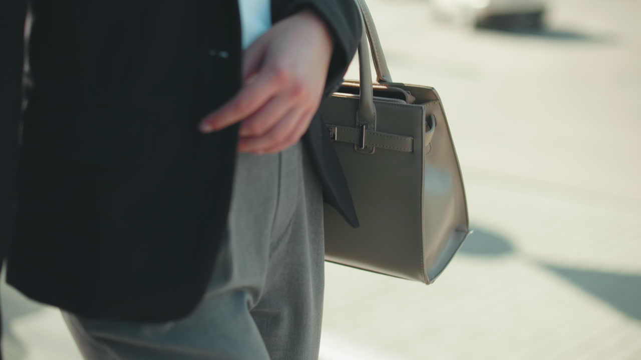 Close up partial view of lady in business outfit carrying handbag walking by on city sidewalk, with blur view of moving car in background