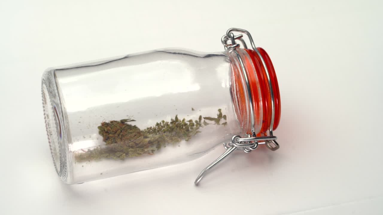 A slow pan reveal of a clear jar containing crushed marijuana lying on its side on a white table