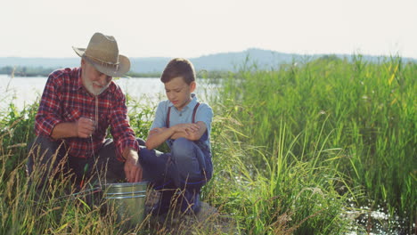 senior fisherman in a hat with his grandson sitting on the shore of the lake and putting a fish in the backet