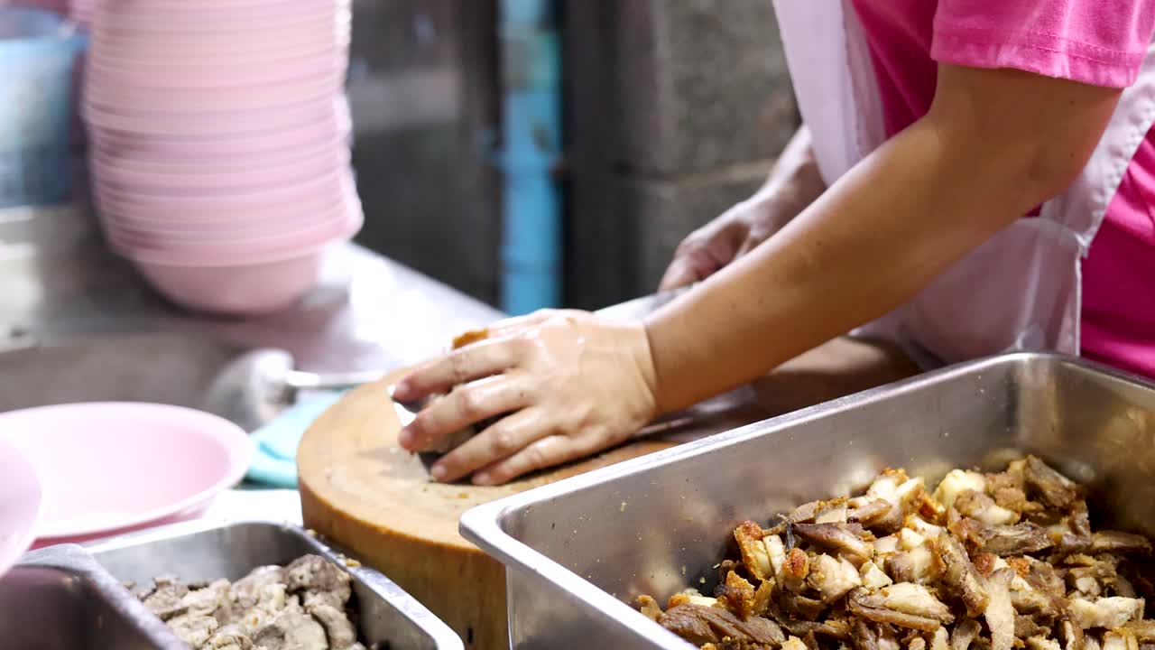 Penjual dengan celemek merah muda mengiris perut babi renyah di bawah pencahayaan pasar makanan kaki lima yang semarak