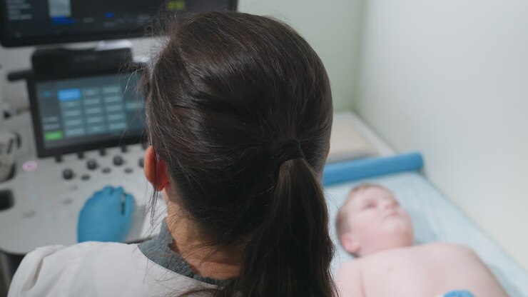 Aerial view of lab scientist with dark ponytail operating ultrasound machine while focused on screen displaying diagnostic data as barechested boy lies faintly in background on patient bed