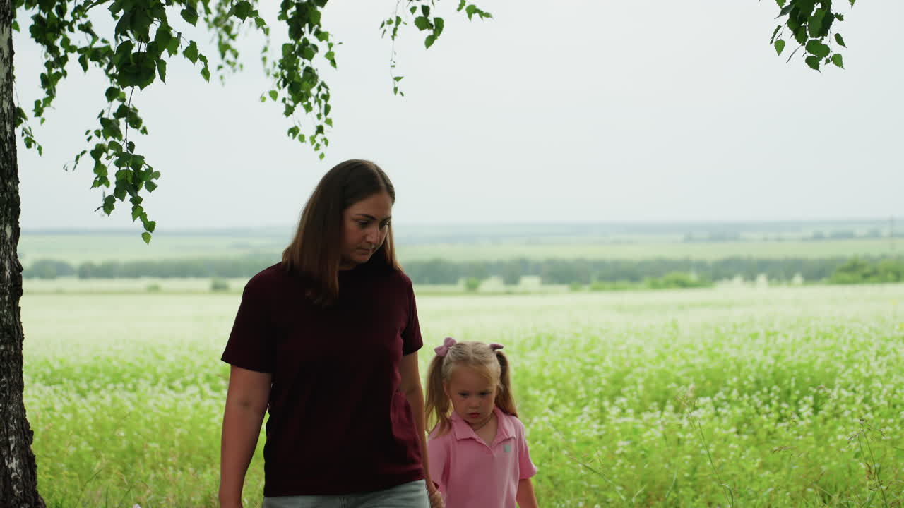madre e figlia bianche che camminano sotto una betulla, scena tranquilla e riflessiva con tono protettivo, la madre osserva la figlia, passi lenti e costanti lungo un pendio erboso, cielo nuvoloso, panorama verde e calmo, delicato