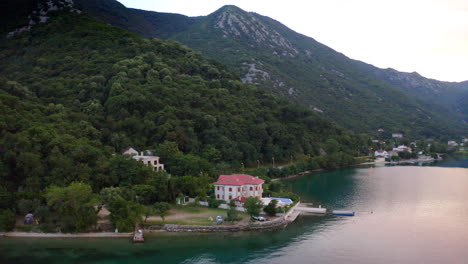 a luxurious villa with a swimming pool and a stone pier on the shores of kotor bay in montenegro, surrounded by trees and a steep mountainside,light evening glow over the area and the bay waters