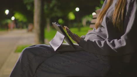 close-up of young girl sitting on park bench at night, focused on tablet under soft park lighting, her hand gestures as she interacts, while people stroll in blurred background