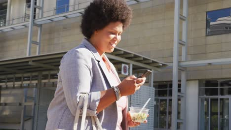 mujer biracial feliz de talla plus caminando con teléfono inteligente y ensalada en la ciudad