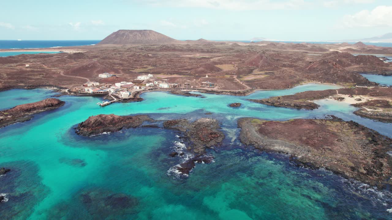 Turquoise waters, rocky coastlines, and volcanic terrain with distant mountains, showcasing coastal beauty and serene landscapes. 4k aerial drone view of Puertito Isla de Lobos, Fuerteventura