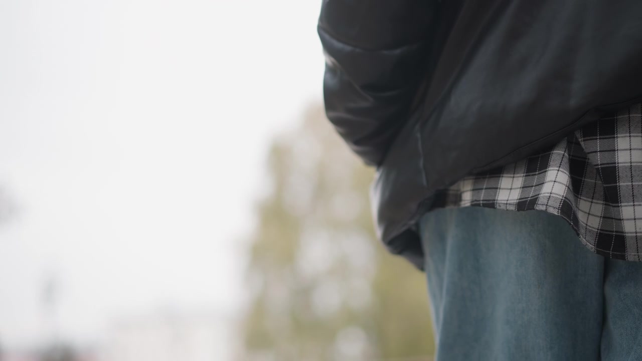 Primo piano di una persona che indossa jeans blu e abbigliamento a strati, con tessuto scozzese visibile sotto il capospalla. Il soggetto rimane immobile con movimenti minimi, concentrandosi sulla zona del busto inferiore.