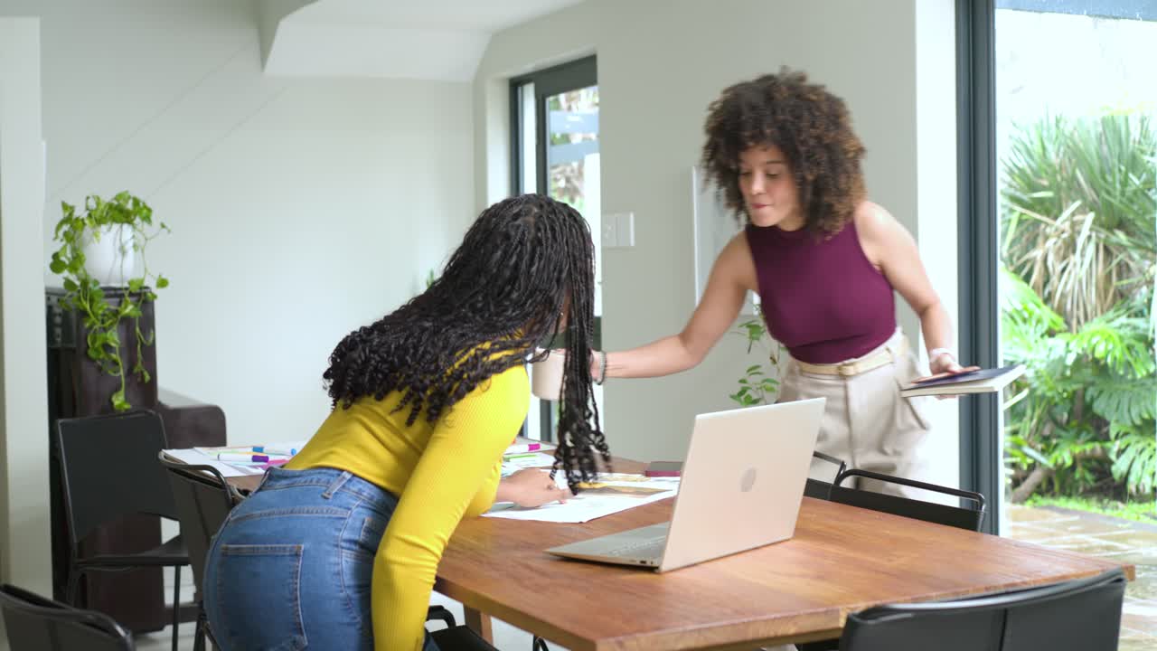 Une femme tenant un ordinateur portable et faisant un geste vers l’écran explique ses idées à des collègues de sexe féminin qui trient des impressions à un bureau.