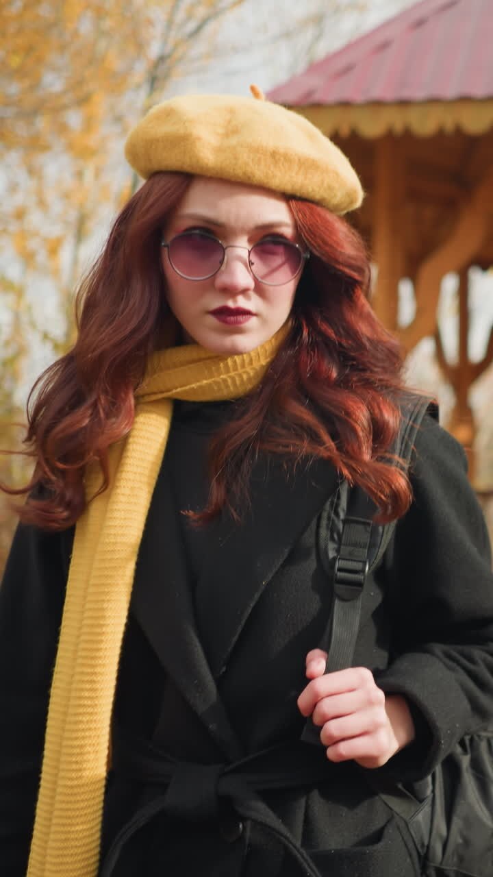 Signora con cappotto nero, sciarpa gialla e berretto che esce da un gazebo di legno in un sereno parco autunnale, sistemando la tracolla della borsa, le calde tonalità dorate degli alberi completano l'ambiente rustico.
