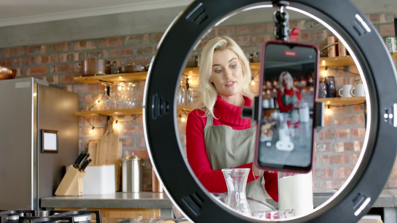 Dando un'occhiata allo smartphone illuminato da ring light, una donna con il grembiule versa il contenuto di un barattolo in una brocca nella cucina di casa per un tutorial