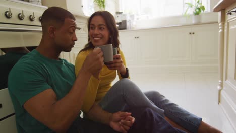 Glückliches-Gemischtrassiges-Paar,-Das-Auf-Dem-Küchenboden-Sitzt,-Kaffee-Trinkt-Und-Redet