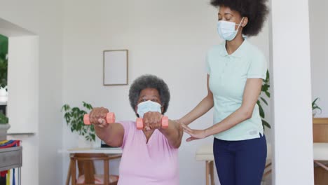 fisioterapeuta afroamericana que usa una máscara facial ayudando a una paciente anciana a hacer ejercicio