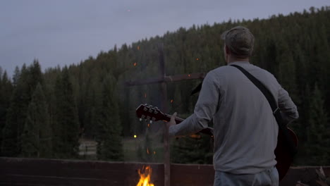 man playing worship music with a guitar next to a campfire and a wooden cross on a full moon evening in the woods of colorado, slow motion tilt down