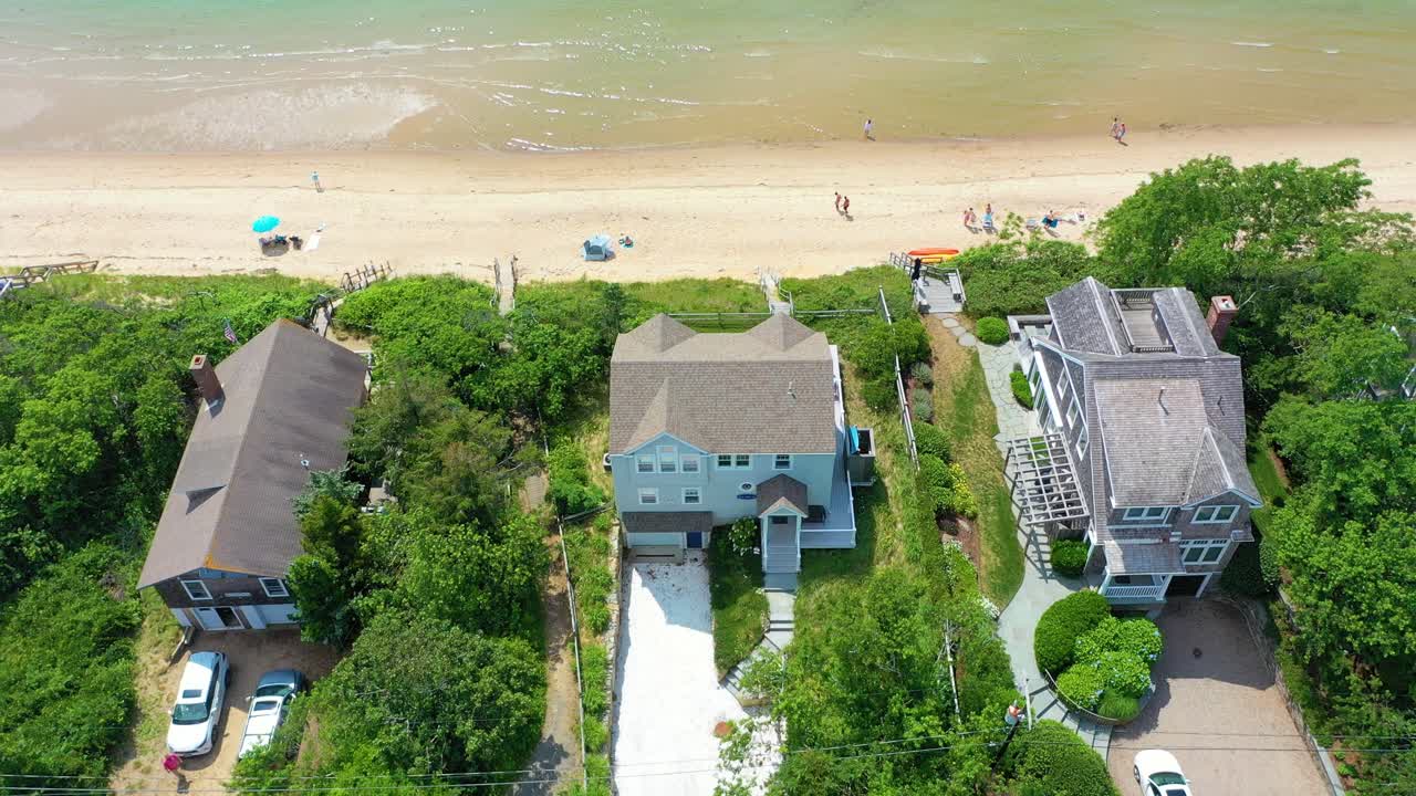Drone orbit of a beachfront home with sandy shoreline stretching outward, waves shimmering in bright summer light, and families scattered along the beach below