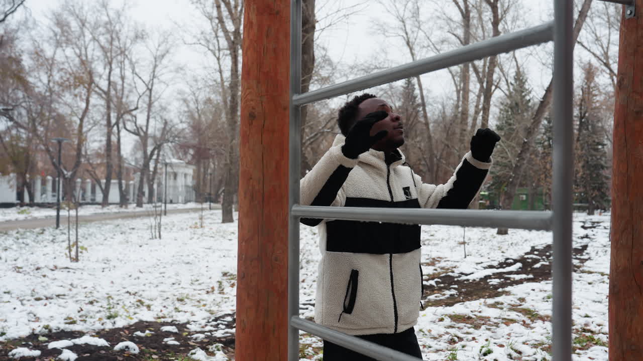남자는 눈 인 공원에서 야외 운동 장비에 오르기 위해 준비하고, 배경에 벌거벗은 나무와 함께 추진 운동을 준비하고, 추운 날씨에 결단력을 강조합니다.