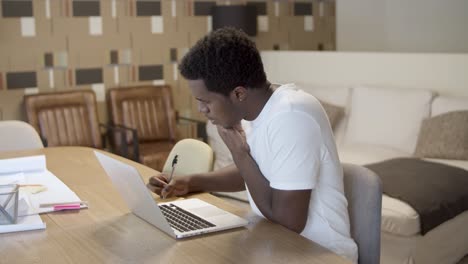 gerente afroamericano hablando por teléfono celular, revisando la computadora portátil y escribiendo en una nota adhesiva