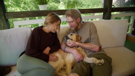 A-blonde-girl-in-a-red-sweater-and-a-blonde-guy-in-glasses-with-a-beard-in-a-light-brown-T-shirt-are-stroking-their-light-colored-dog-on-a-sofa-in-nature-in-the-gazebo