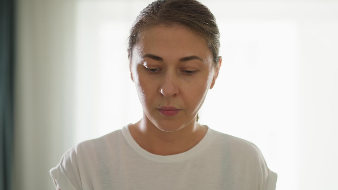 Woman gazes downward against bright backdrop closeup. Focused lady in white top stands near window in light-colored apartment. Attractive woman face