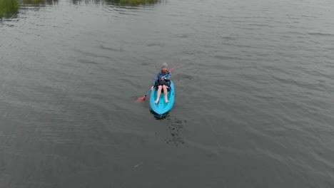 órbita aérea alrededor de un hombre barbudo de jengibre remando en un kayak azul entre un lago pantanoso en australia