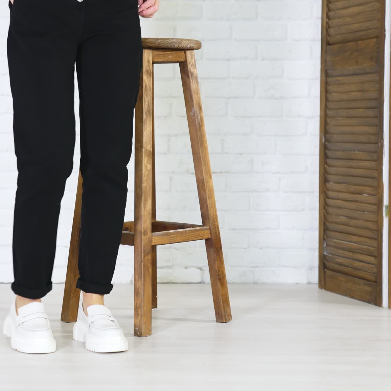 Woman in black jeans comes to a wooden stool showing white shoes. Footwear presentation at white backdrop