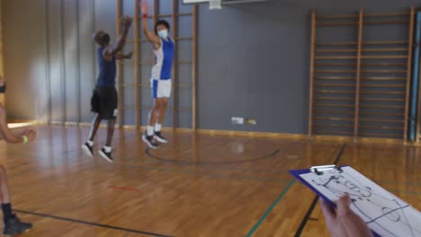 equipo de baloncesto masculino diverso jugando un partido con el entrenador sosteniendo el clipboard, usando máscaras faciales