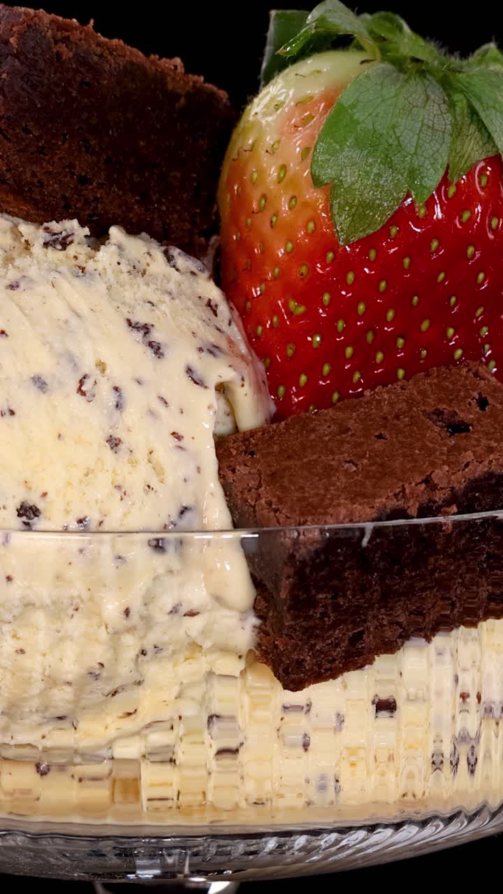 Una mano coloca una fresa fresca y trozos de brownie sobre una bola de helado de vainilla con trocitos de chocolate en un tazón de vidrio bajo una brillante iluminación de estudio.