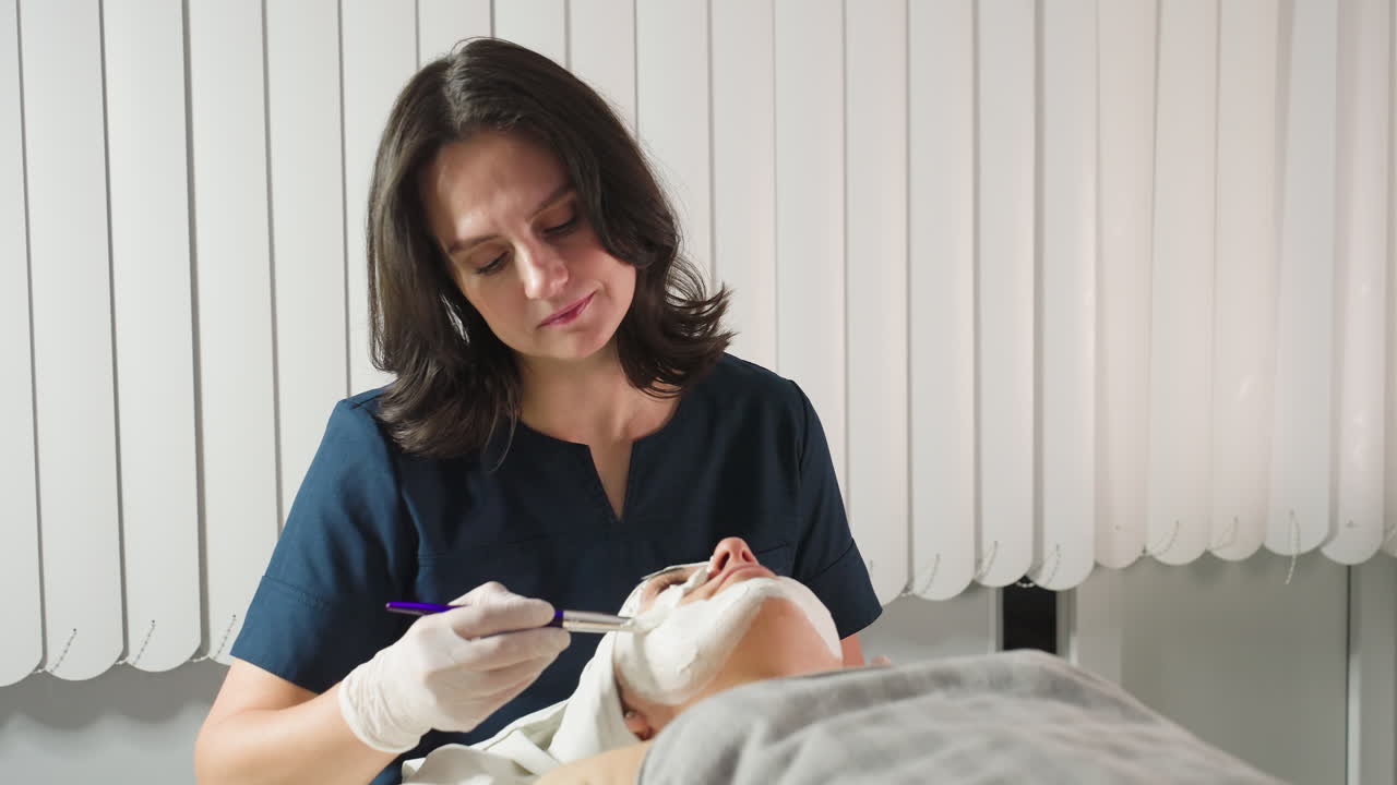 Wellness Technician Applies Face Scrub Carefully With Soft Silicone Brush On Client Skin During Relaxing Spa Treatment Under Bright Studio Light Emphasizing Gentle Technique And Professional Care