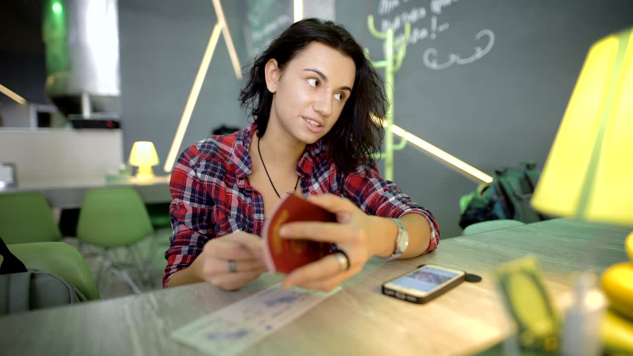 joven con cabello negro corto, vestida con una camisa en un reloj de mano, sentada en la barra en la cafetería del aeropuerto, esperando su salida, considerando su pasaporte, y se comunica con alguien