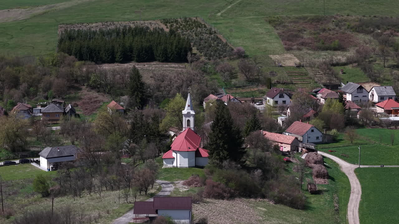 Eine Luftaufnahme zeigt ein charmantes Dorf, dessen markante Kirche sich zwischen traditionellen Häusern erhebt. Die Siedlung liegt eingebettet in grüne sanfte Hügel und stellt ein friedliches europäisches Landleben dar.