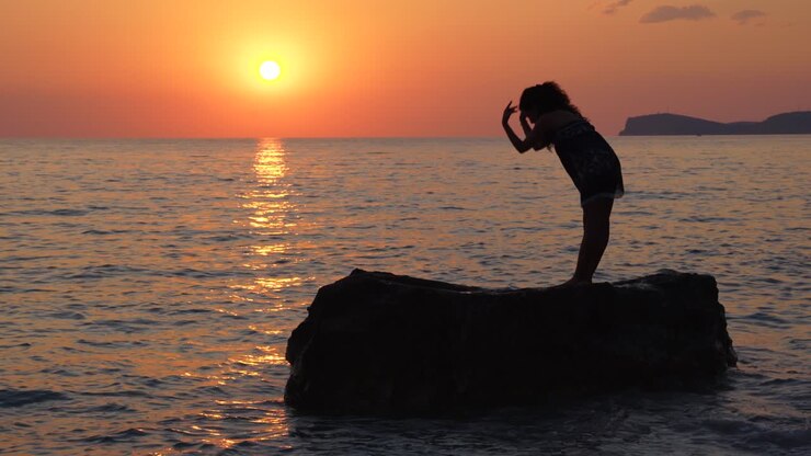 Giovane donna esegue esercizi di concentrazione yoga in piedi su una scogliera bagnata dall'acqua del mare al bellissimo tramonto.