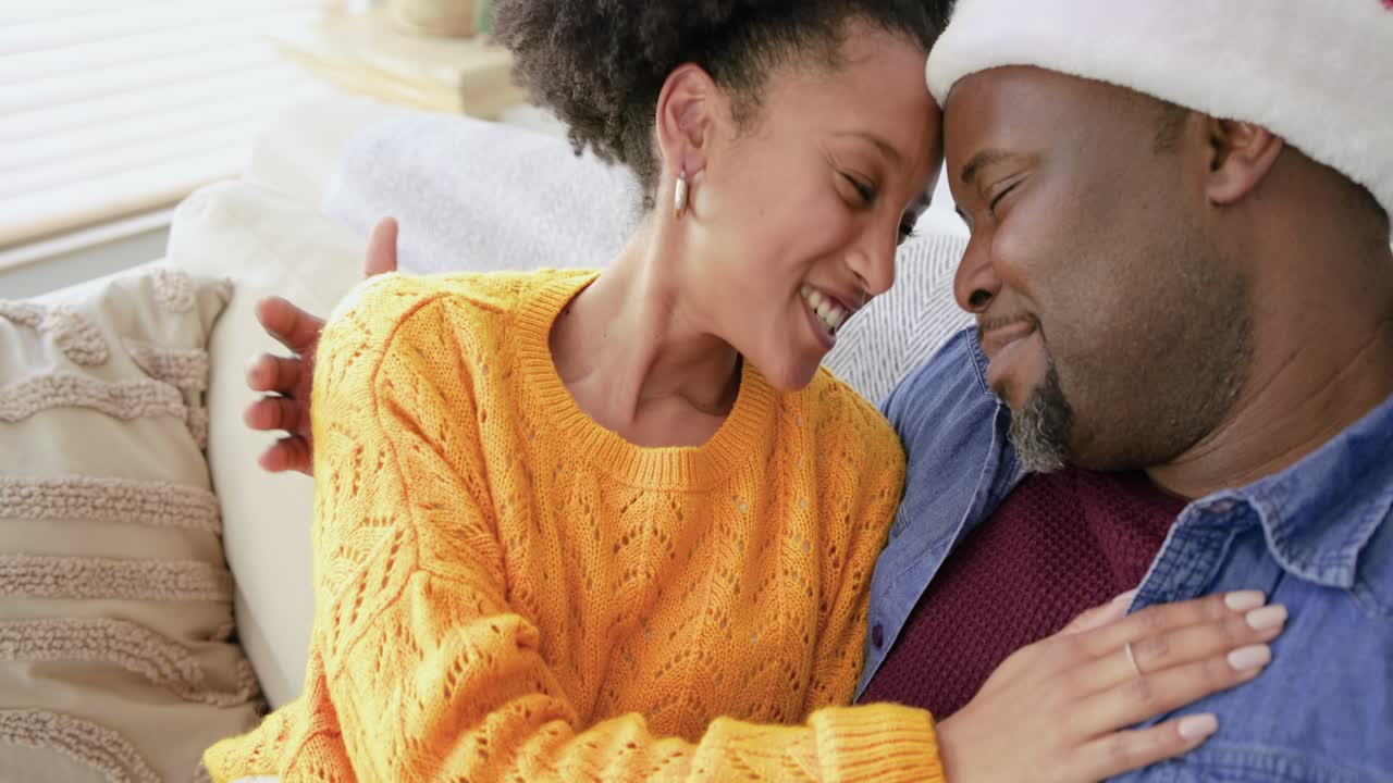 Mid adult couple wearing Santa hat exchanging glances, nestling under knit blanket on sofa at home