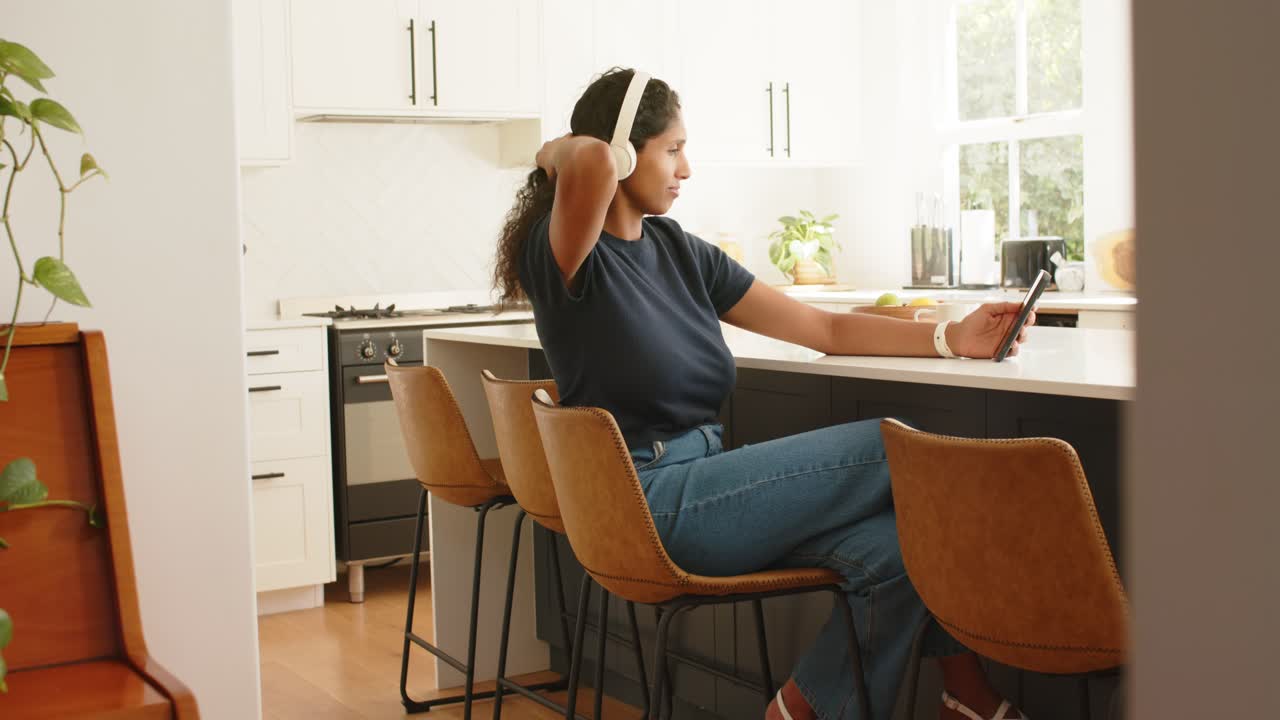 African American woman sitting at kitchen island answering phone alert, adjusting hair, headphones