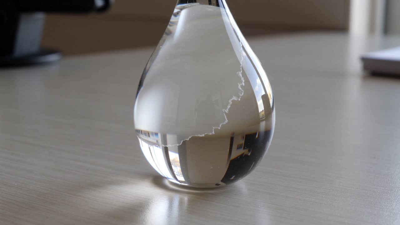Cristalli che si muovono e reagiscono alla luce all'interno di un vetro a forma di goccia sulla scrivania, con un dispositivo sottile e un oggetto scuro.