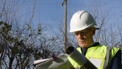 Low-angle-portrait-of-an-architect-building-inspector-inspecting-a-construction-site-with-a-clip-board-and-architectural-plans