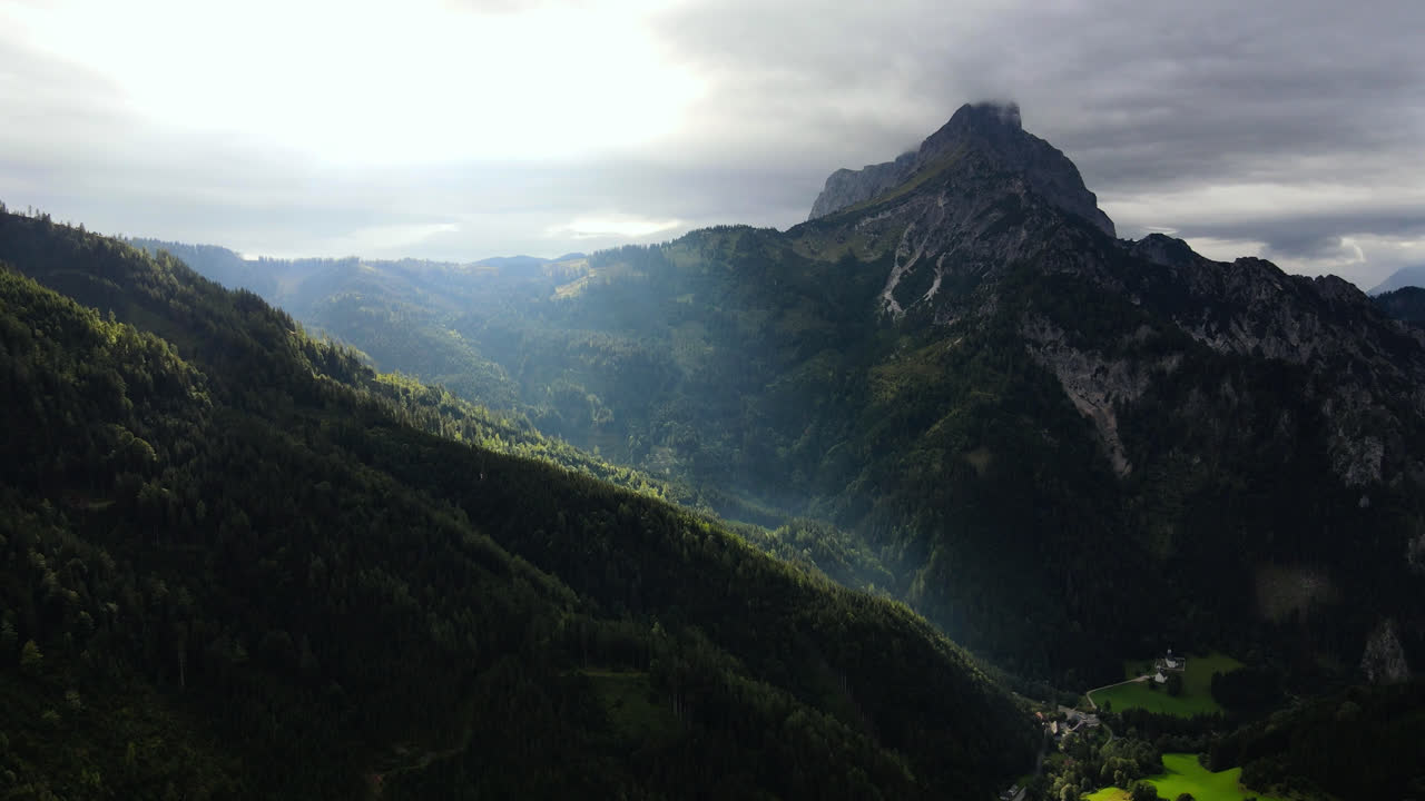 una toma lenta de un dron de un pico de montaña sobre johnsbach en el parque nacional de gesause, austria con el sol brillando a través de las nubes