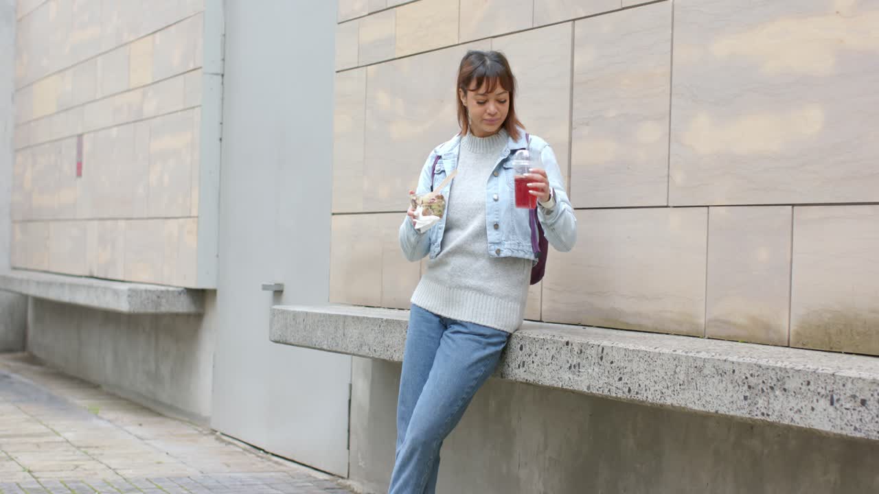 Donna asiatica adulta appoggiata a una panchina, che sorseggia una bevanda, osserva il camminamento e addenta cibo da asporto. Spazio per la copia.