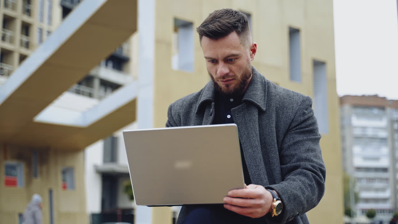 Portrait of a handsome man with a laptop. Young bearded businessman in a coat works on a wireless gadget on the urban background outdoors.