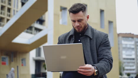 portrait of a handsome man with a laptop. young bearded businessman in a coat works on a wireless gadget on the urban background outdoors.